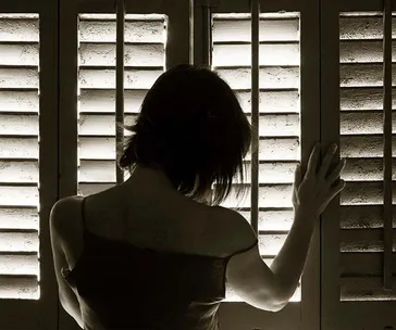 A woman stands pensively at window blinds, her hand touching the slats, silhouetted by light filtering through.