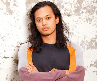 Khanh Ong with long hair, crossed arms, wearing a colorful sweater against a textured wall.