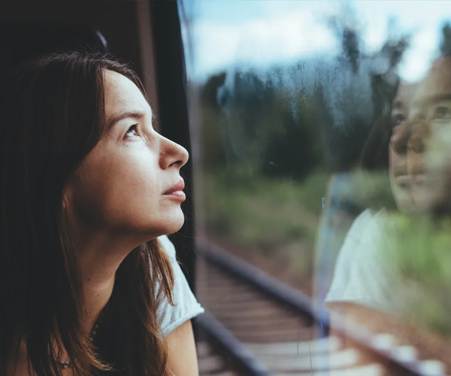 Woman gazing out a train window with her reflection visible in the glass.