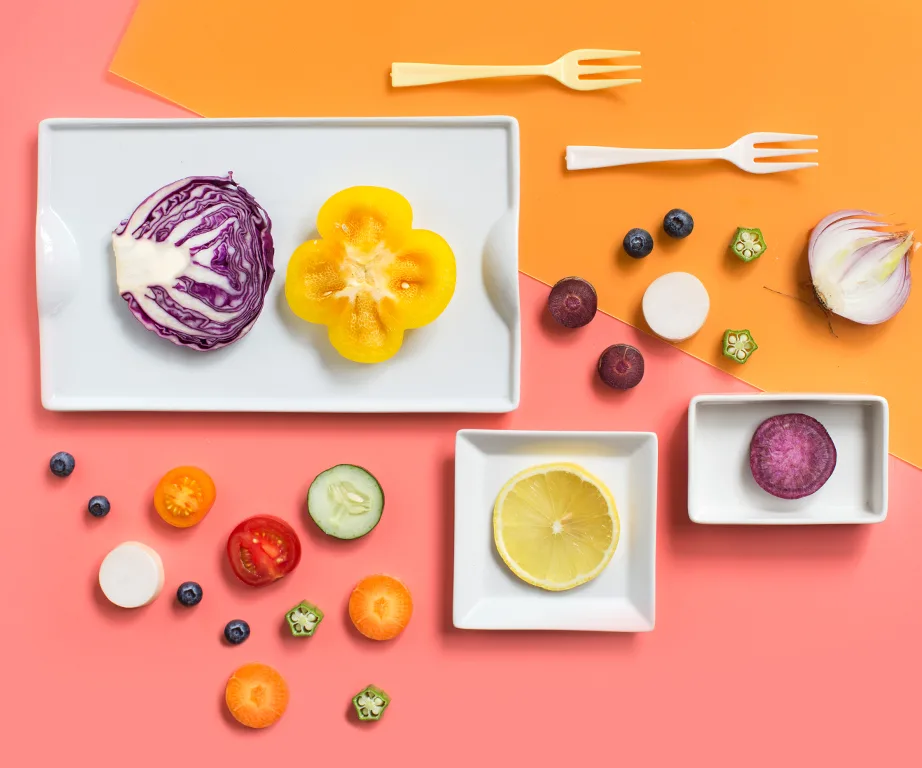 Assorted colorful fruits and vegetables on two-tone background with small white trays and forks.