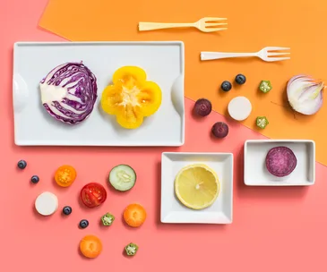 Assorted colorful fruits and vegetables on two-tone background with small white trays and forks.