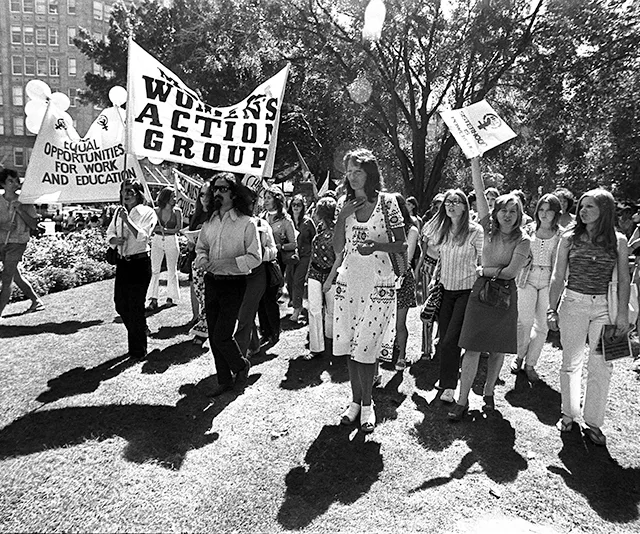 Protesters march with "Women's Action Group" and "Equal Opportunities" signs in black and white photo.