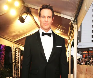 A man in a tuxedo under a canopy walkway at an event, with bright lights in the background.