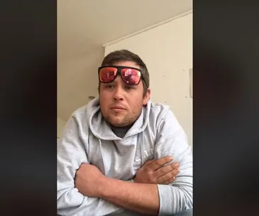 A man in a hoodie with sunglasses on his head, arms crossed, looking serious and contemplative.