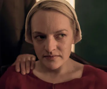 A woman wearing a bonnet looks to the side with a hand resting on her shoulder, a scene from The Handmaid's Tale.