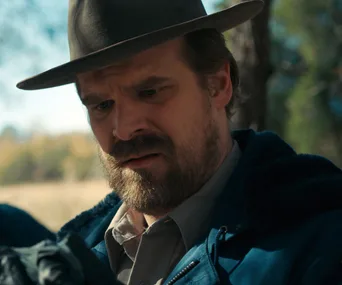 Man in a hat and jacket looking down thoughtfully in an outdoor setting.