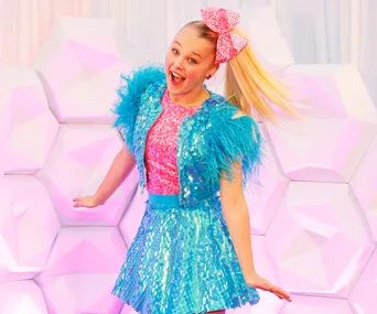 Joyful person in vibrant blue and pink outfit with sequins and a large bow, posing against a geometric pastel backdrop.