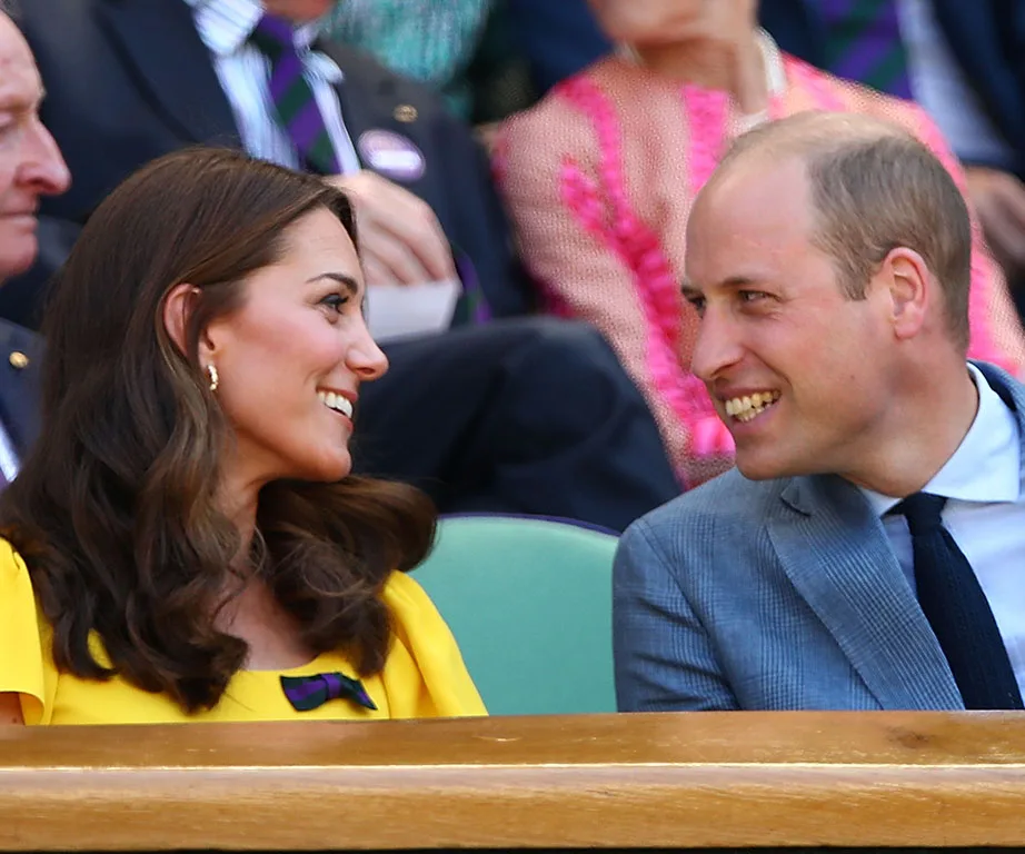 A couple smiling at each other in colorful attire, seated at an outdoor event with people in background.