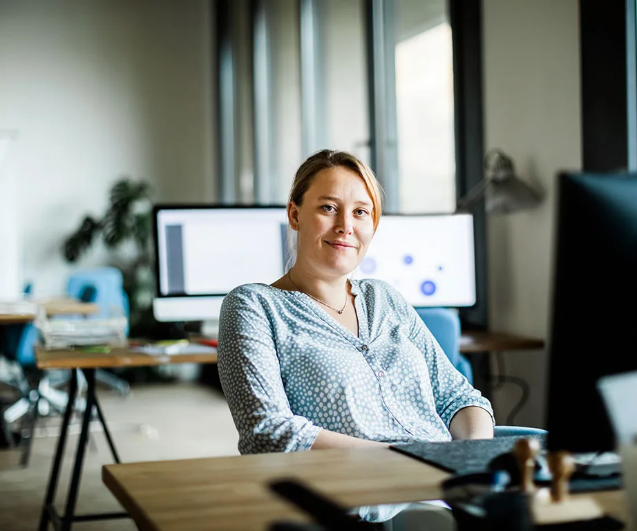 Person sitting in a modern office, smiling, with computer screens and desks visible in the background.