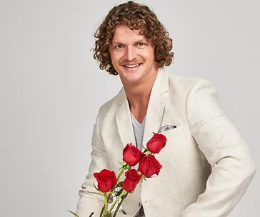 Man in a beige jacket holding a bouquet of red roses, smiling at the camera.