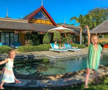 Children playing by a luxurious pool villa with parents relaxing on sun loungers under a large umbrella.