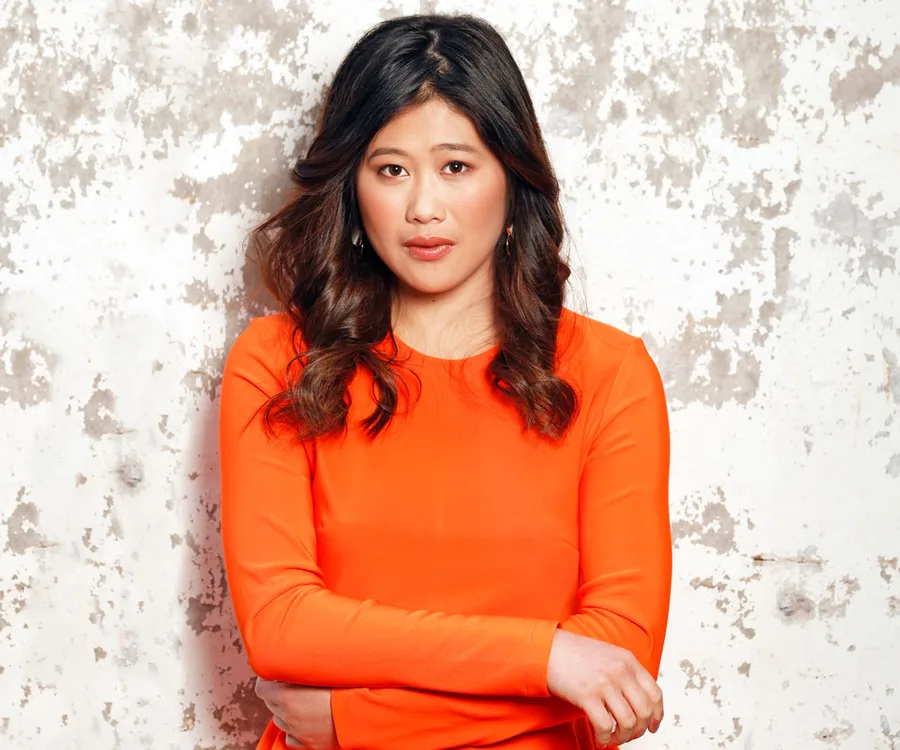 A woman in an orange top stands against a textured white wall, with arms crossed and a neutral expression.