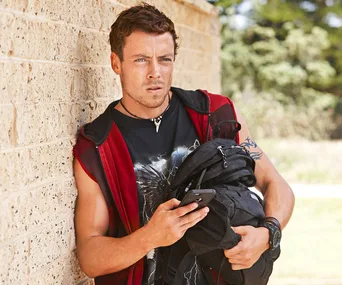 A man leans against a brick wall, holding a backpack and looking at a phone, wearing a red vest and a necklace.