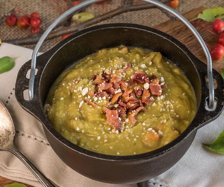 A black pot filled with pea and ham soup, topped with crispy bacon bits and grated cheese.