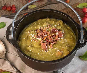 A black pot filled with pea and ham soup, topped with crispy bacon bits and grated cheese.
