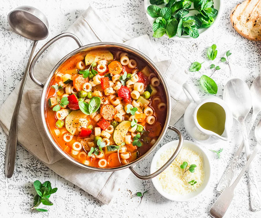 A pot of colorful minestrone soup with vegetables and pasta, garnished with herbs, surrounded by olive oil, cheese, and greens.
