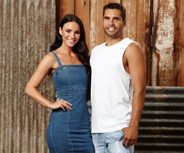 A woman in a denim dress stands beside a man in a white tank top, both smiling against a rustic wooden and corrugated metal backdrop.