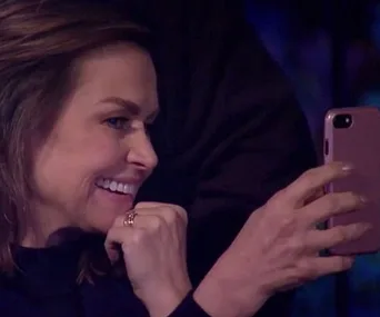 Woman smiling, holding a phone horizontally, appearing to take a photo or video in a dimly lit setting.