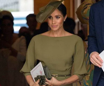 Meghan Markle wearing an olive green dress and hat, holding a program, at Prince Louis' christening event.