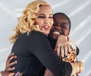 A woman with blonde hair hugs a smiling child against a light background.