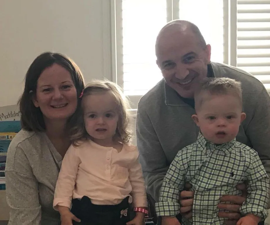 Family photo of smiling parents with young son and daughter indoors, with light streaming through the window blinds.