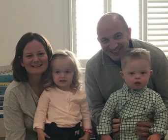Family photo of smiling parents with young son and daughter indoors, with light streaming through the window blinds.