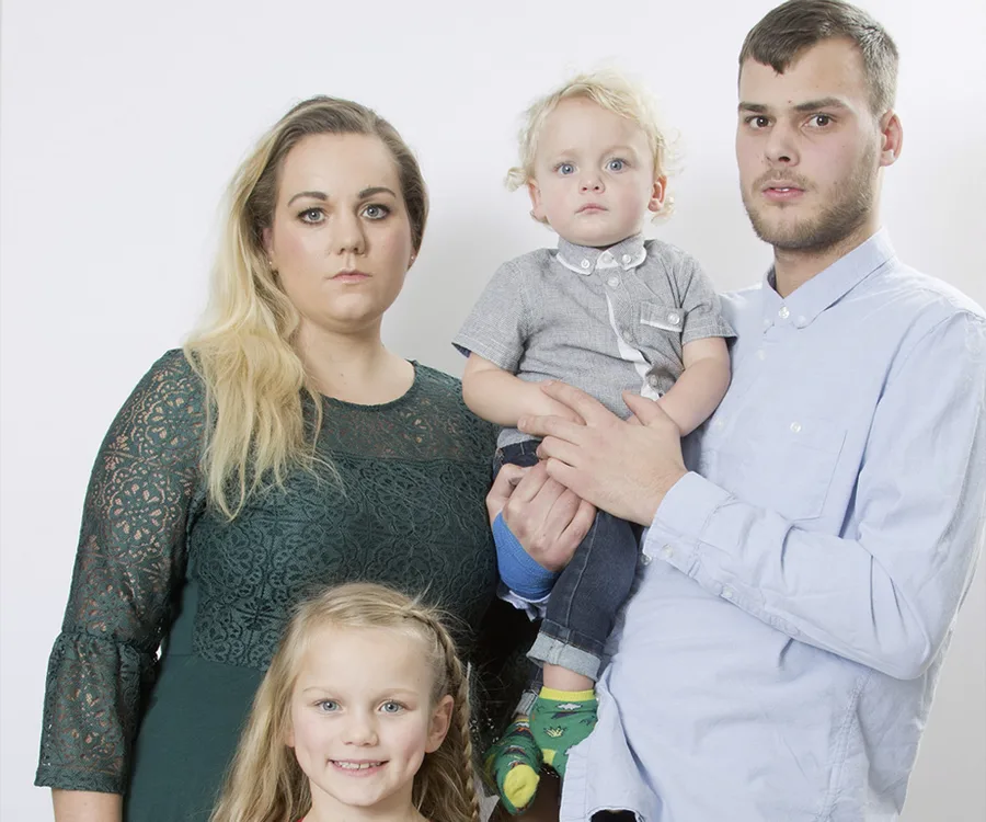 A family of four posing together; parents each holding a child, with a neutral background.
