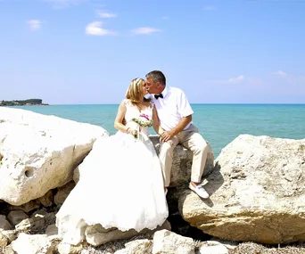 A bride and groom kissing on rocks by a beach, with a turquoise ocean and clear blue sky in the background.