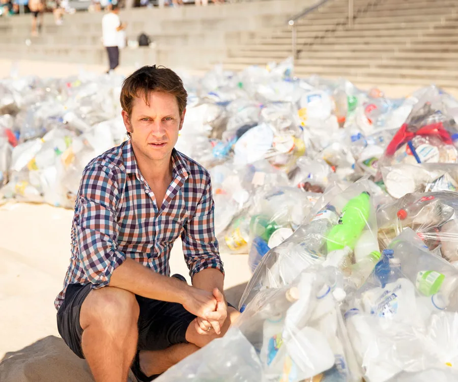 A man in a checkered shirt kneels beside numerous bags of plastic waste on a beach with steps in the background.
