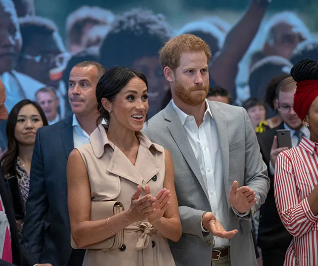 Prince Harry and Meghan Markle at Nelson Mandela exhibition, smiling and clapping among a crowd.