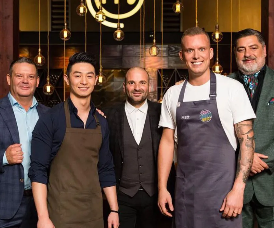 Group of five men smiling, two wearing aprons with "MasterChef" branding, standing in a restaurant setting with warm lighting.