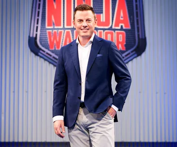 Man in a blue blazer standing in front of a "Ninja Warrior" sign on a corrugated metal background.