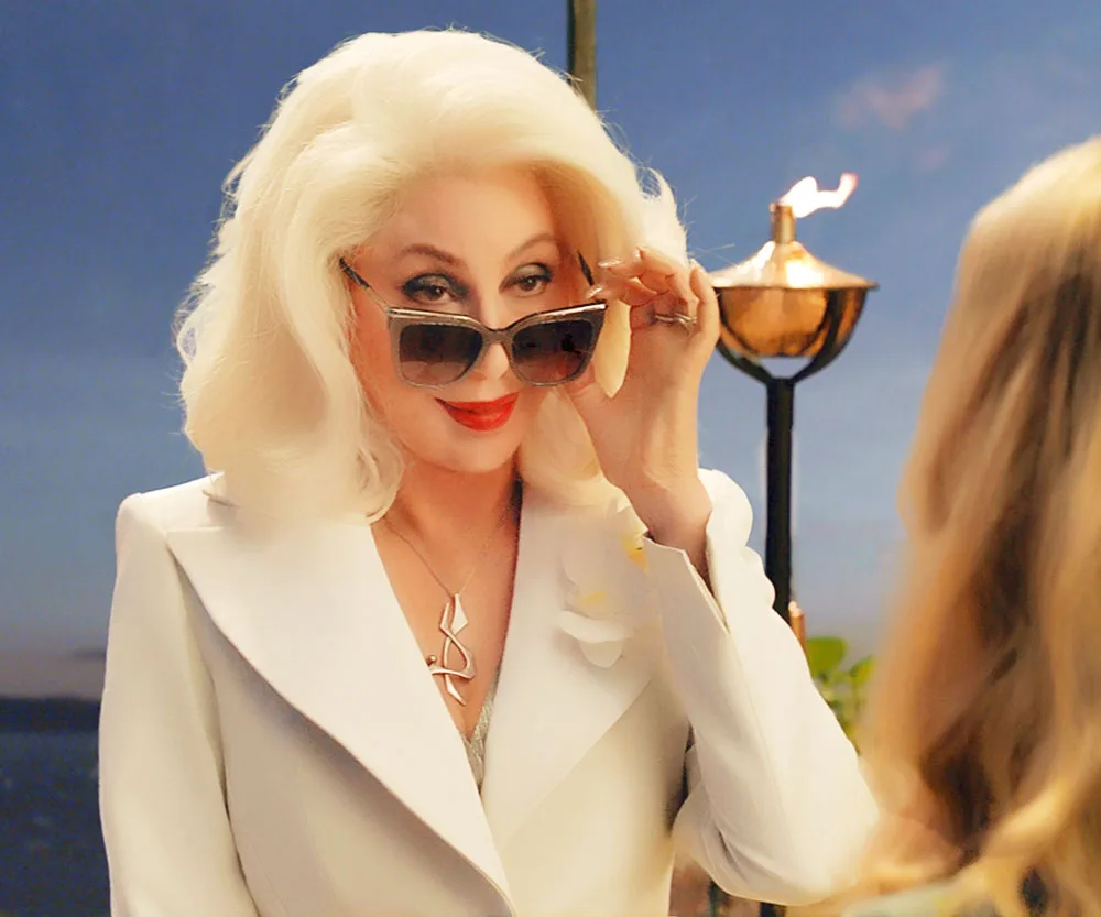Cher in a white suit lowers sunglasses in a scene from "Mamma Mia! Here We Go Again" with a scenic background.