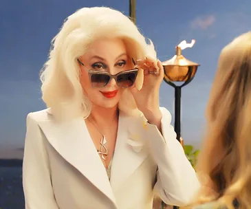 Cher in a white suit lowers sunglasses in a scene from "Mamma Mia! Here We Go Again" with a scenic background.