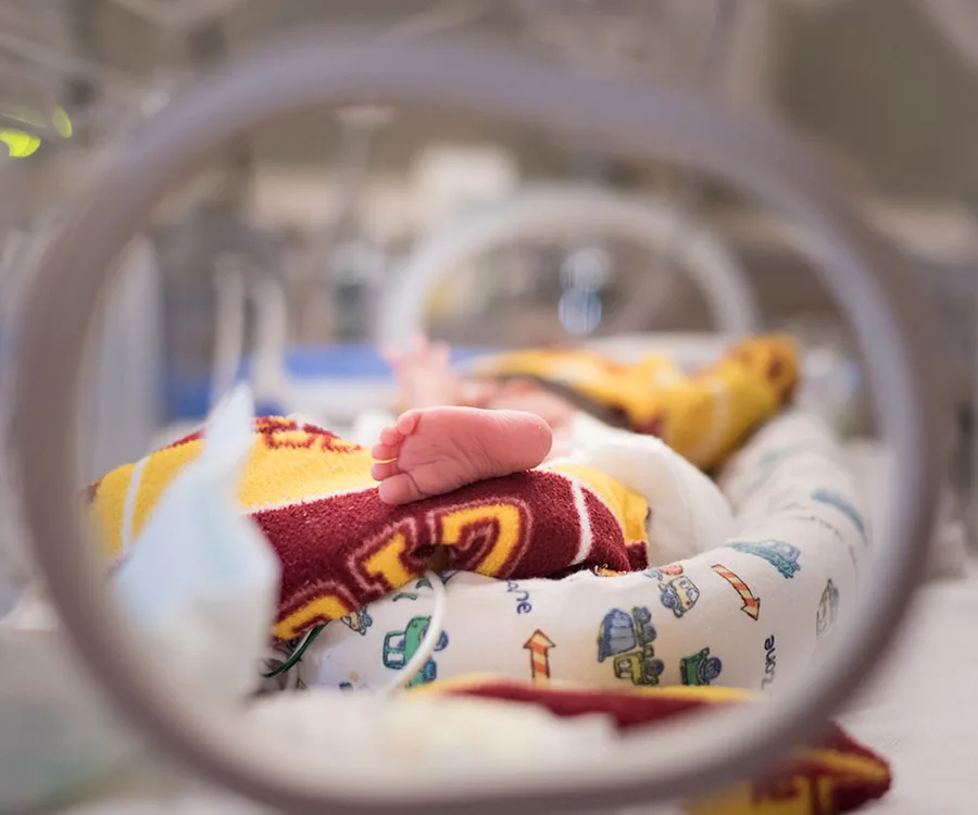 Premature baby in NICU crib, foot visible, covered with colorful blanket.