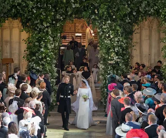 Bride and groom exit church under floral arch, greeted by guests at royal wedding.