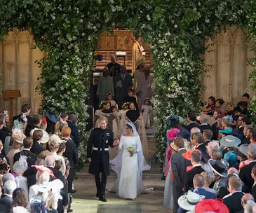 Bride and groom exit church under floral arch, greeted by guests at royal wedding.