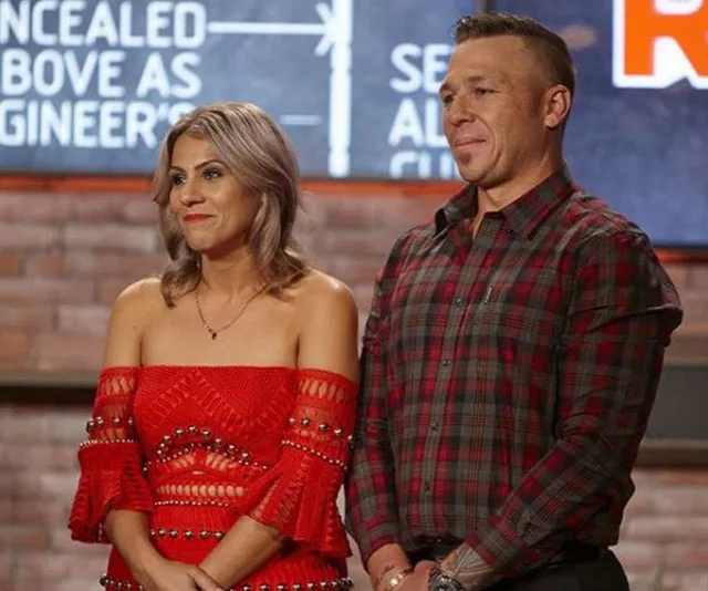 Couple standing on a set, woman in red dress, man in plaid shirt, with text partially visible in the background.