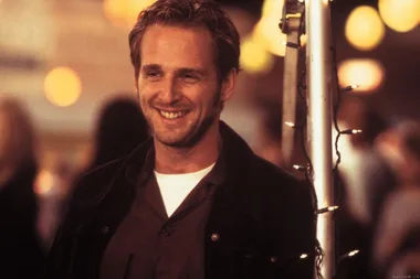 A smiling man with light brown hair stands near string lights, appearing relaxed and happy, possibly at an evening event.