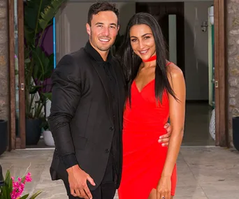 Two people smiling and posing, one in a black suit and the other in a red dress, standing in a stylish indoor setting.