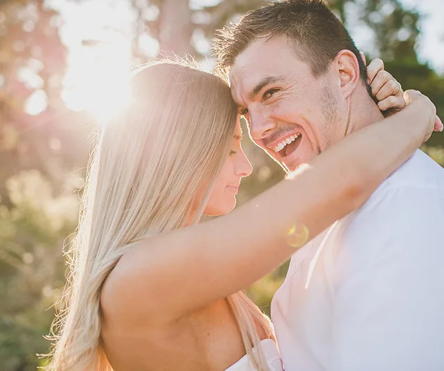 A couple embraces in a sunlit, outdoor setting, both smiling warmly at each other, creating a romantic atmosphere.