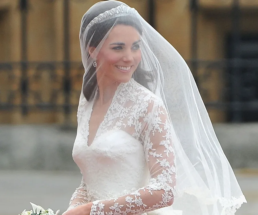 Bride in lace wedding dress with veil and tiara, smiling outdoors.