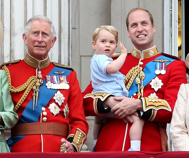 Prince Charles, Prince William, and Prince George in red uniforms on a balcony, with George being held and waving.