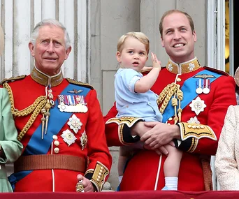 Prince Charles, Prince William, and Prince George in red uniforms on a balcony, with George being held and waving.