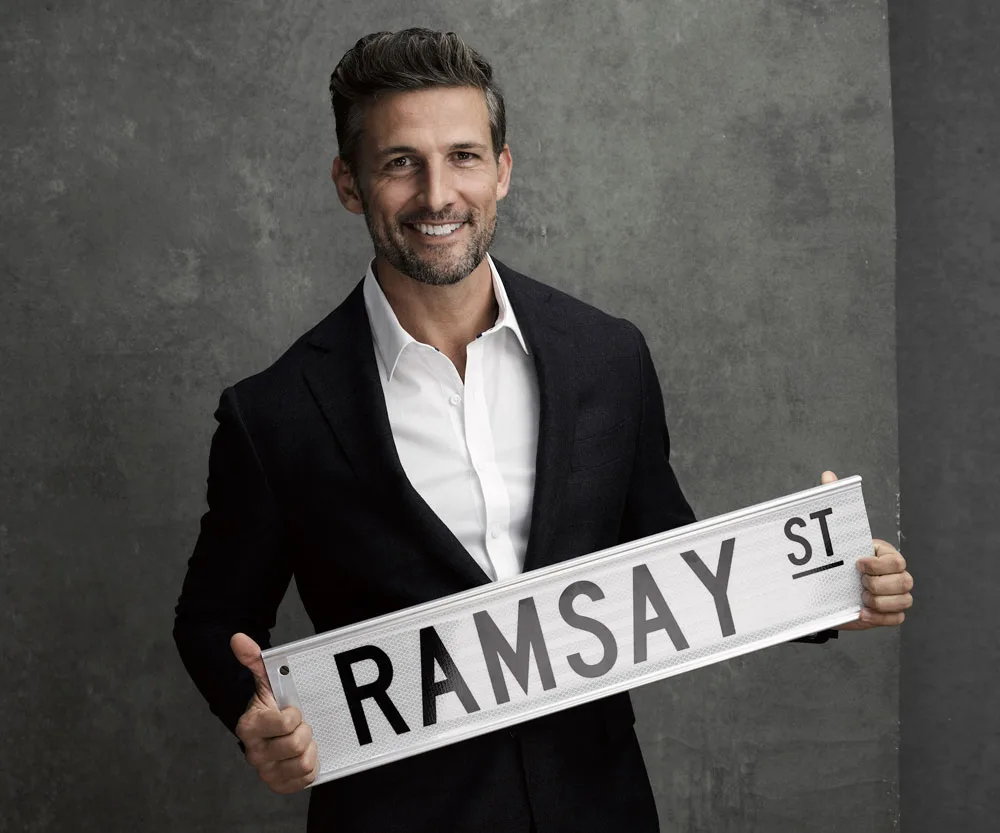 Man in a suit smiling, holding a "RAMSAY ST" sign associated with the TV show "Neighbours."