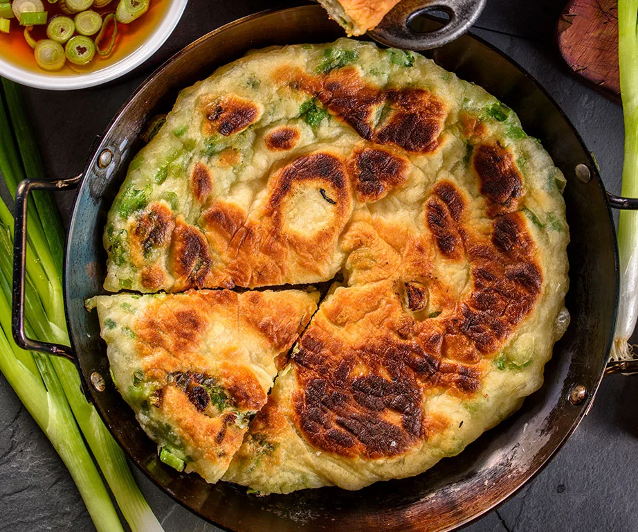 Golden-brown spring onion pancake in a skillet, with dipping sauce and fresh green onions on the side.