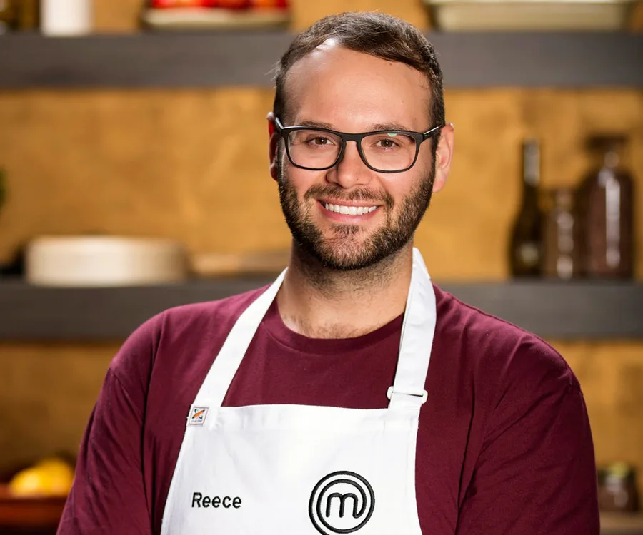 Man with glasses wearing a “MasterChef” apron, smiling, standing in a kitchen setting.