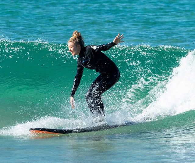 A person in a black wetsuit skillfully surfs a small wave in clear blue water, balancing on a surfboard.