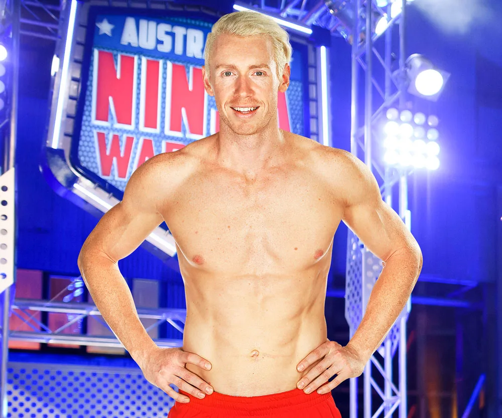 Shirtless athlete on "Australian Ninja Warrior" set with hands on hips, smiling confidently.