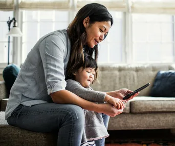 Adult and child sit on sofa, sharing a pleasant moment with a tablet, in a bright living room setting.